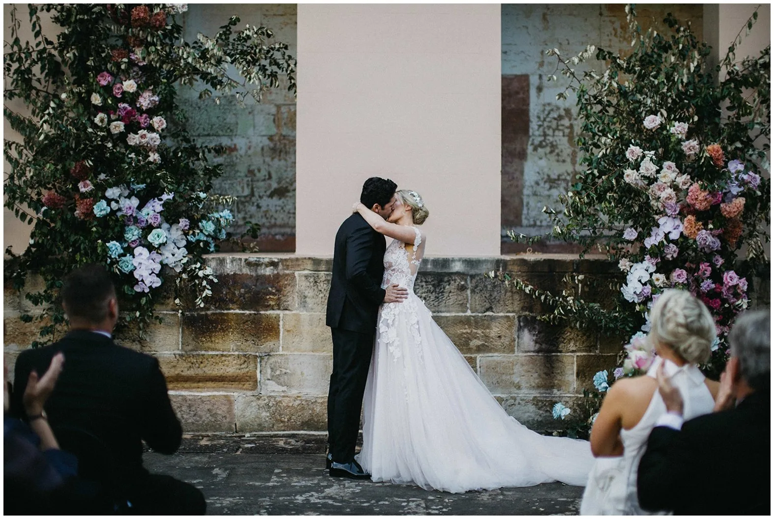 A newlywed couple is kissing at their marriage ceremony at Cowbell Creek wedding venue with guests watching from behind them, photographed by Joshua Mikhaiel