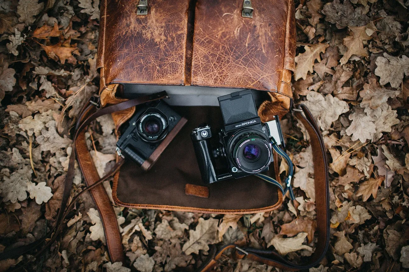 Two film cameras in a worn leather bag resting on autumn leaves